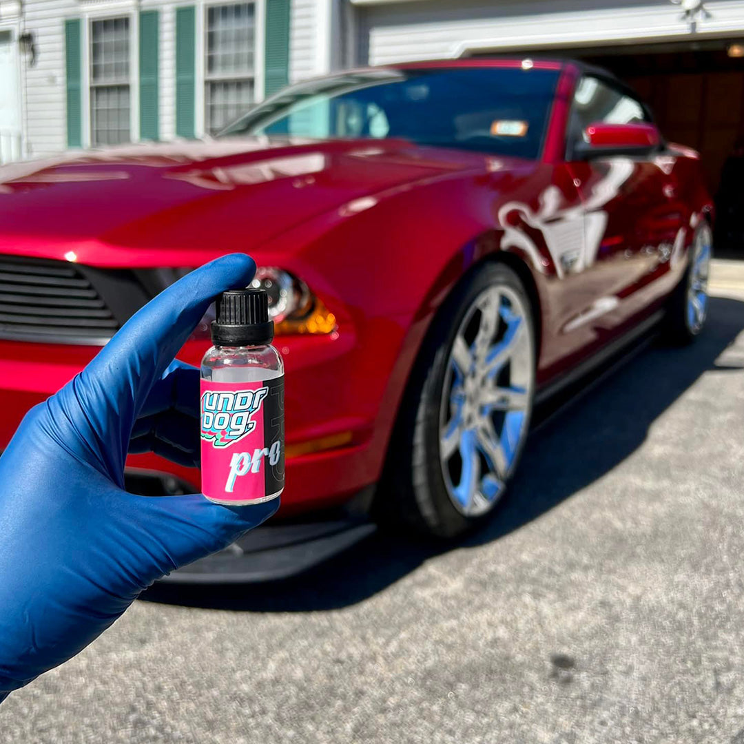 Blue-gloved hand holding an Undrdog Pro bottle in front of a red car