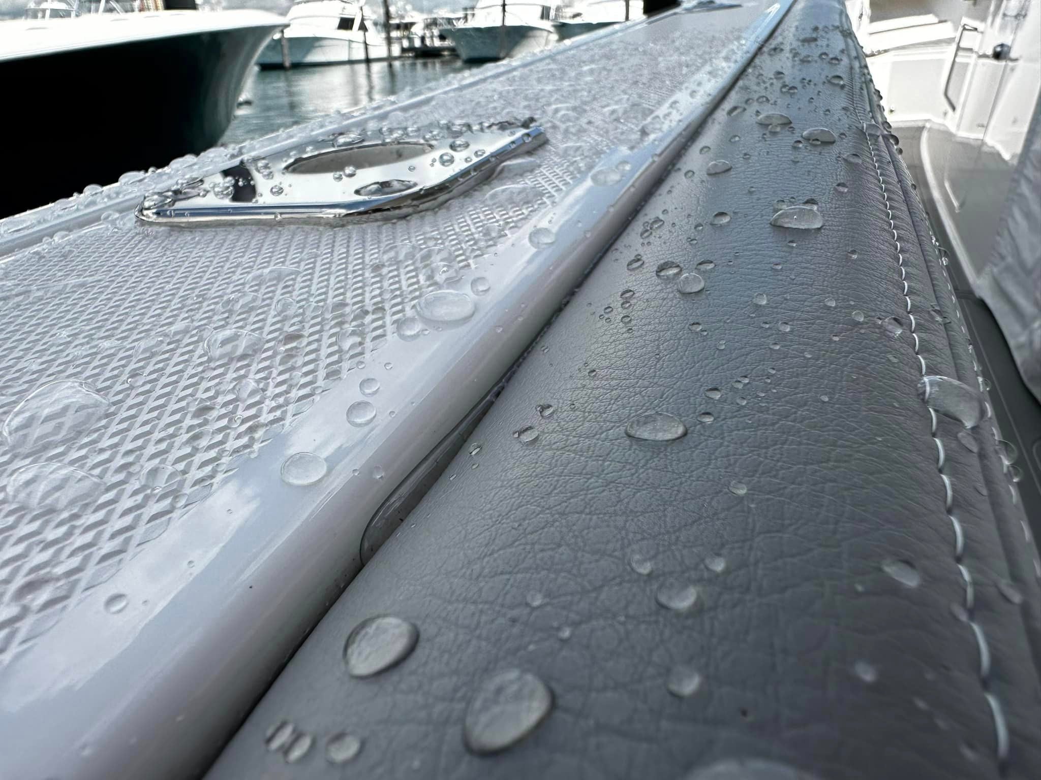 Close-up of water droplets on a textured, light gray surface of a boat, with parts of other boats and water visible in the blurred background at a marina.