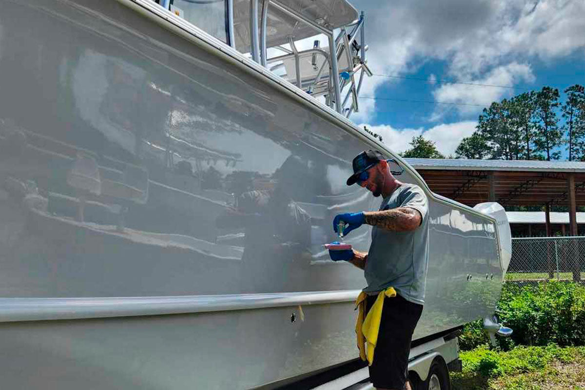 A man wearing a cap and gloves polishes the side of a large white boat using a cloth. The boat is on a trailer, parked outdoors on grass with trees and a building in the background.