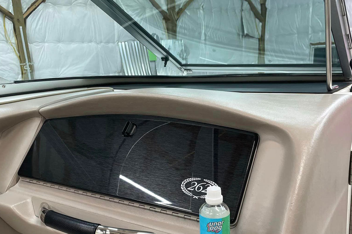 A close-up of a car dashboard with a black glove compartment panel, a “264” sticker, and a bottle of windshield washer fluid labeled “Wind Dog” resting on the beige interior.