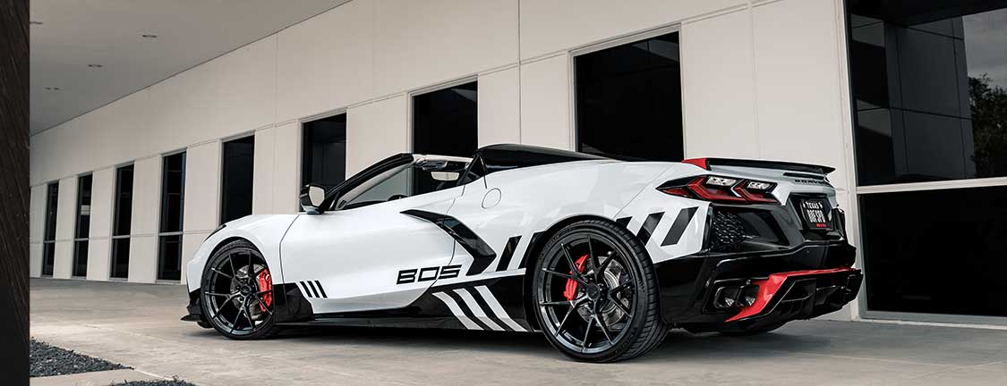 White sports car with black and red accents parked in front of a modern building with large windows. The car has the number "805" on its side and features a sleek design with a convertible top.