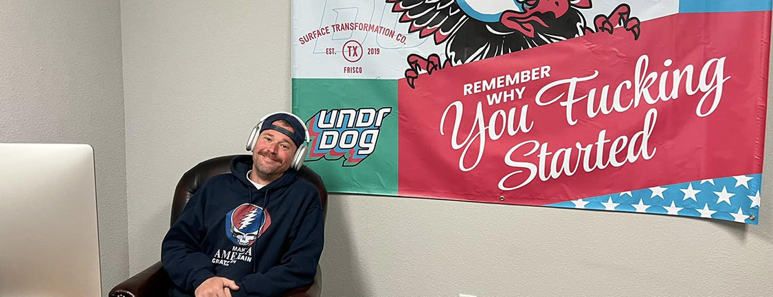 A man wearing headphones and a hoodie sits in a chair next to a desk with an Apple computer. Behind him is a large motivational banner and other signs on the wall.
