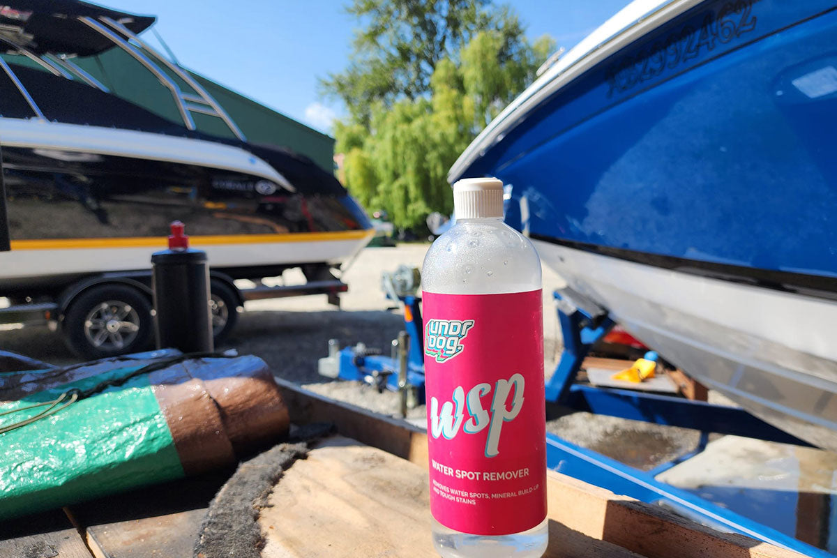A bottle of Underdog Wisp Water Spot Remover sits on a wooden surface outdoors, with two boats and a tree visible in the background.
