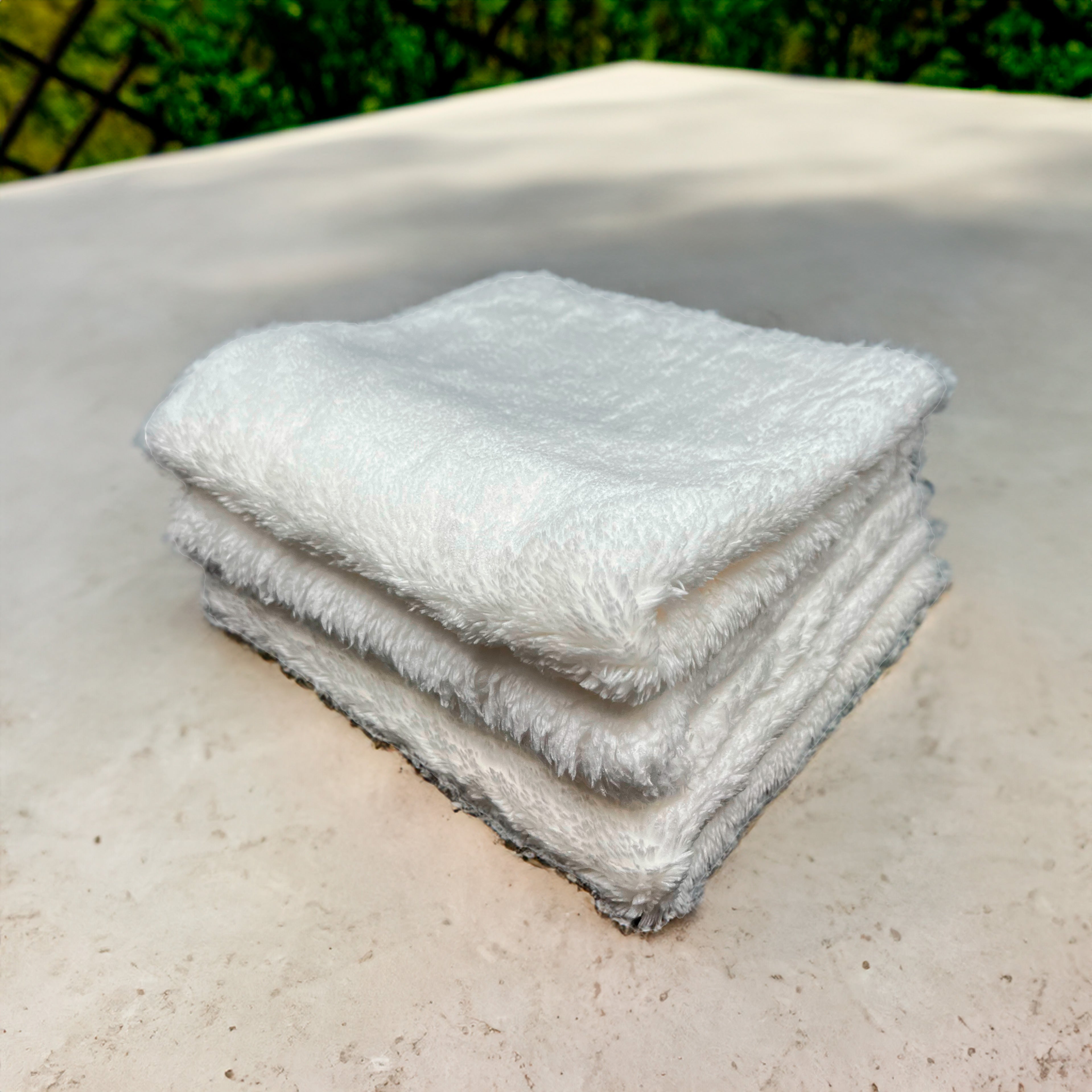 A stack of four neatly folded white towels sits on a light-colored stone surface with a blurred green background and part of a black fence visible.