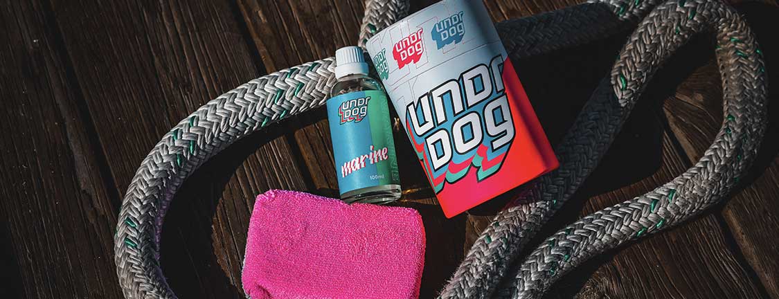A bottle of undrdog Hyper Shine, a container of undrdog protective coating, a red cloth, and a coiled rope are arranged on a wooden surface.