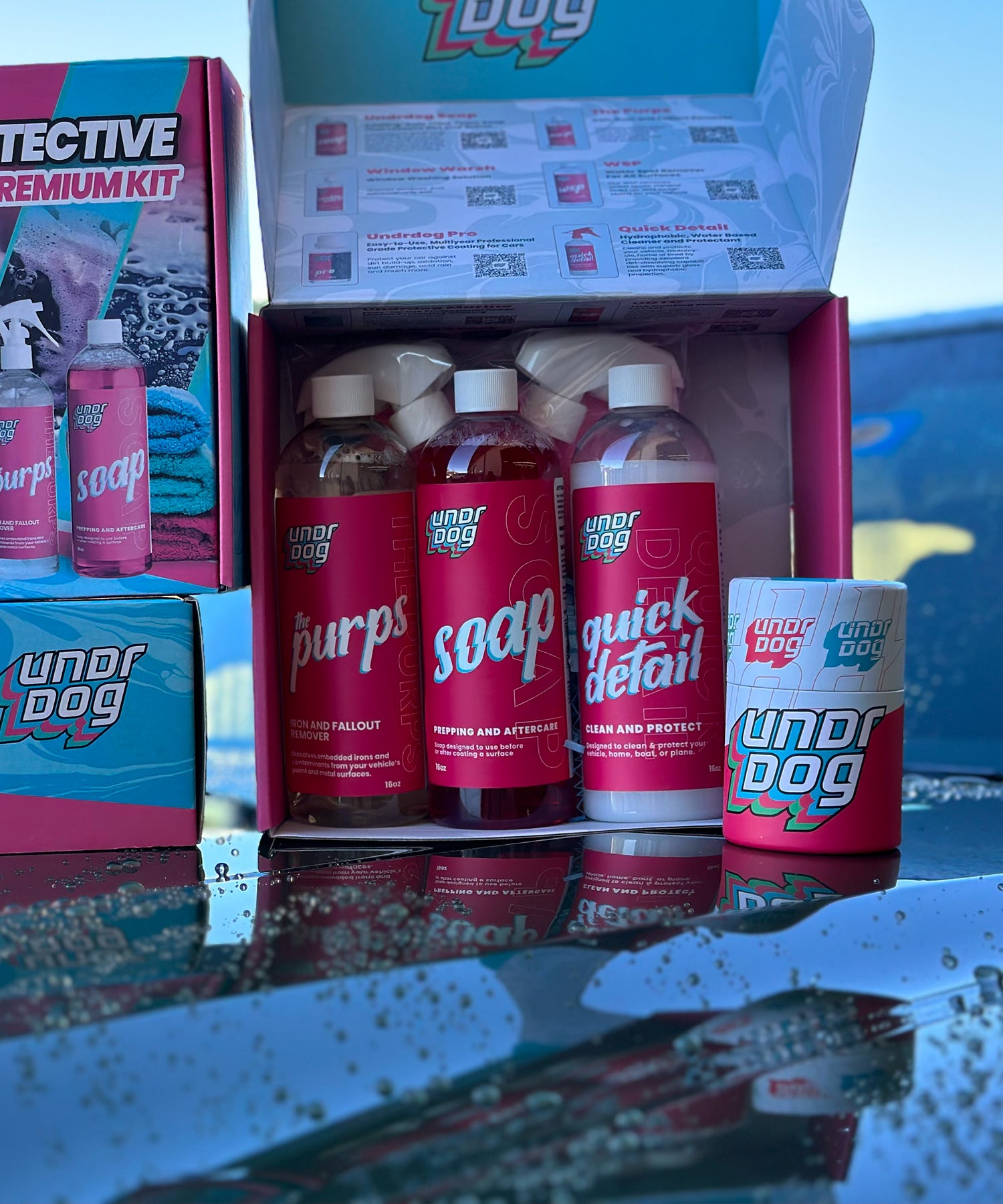 A display of car care products includes multiple bottles labeled "purps," "soap," and "quick detail" with a mug nearby. They are arranged on a shiny surface with water droplets, and the packaging features red and blue branding.