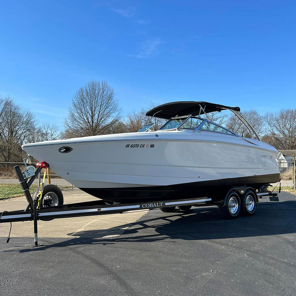 A white Cobalt boat on a black trailer is parked on a paved surface. The boat has a black canopy and visible registration numbers. Trees with bare branches are in the background under a clear blue sky.