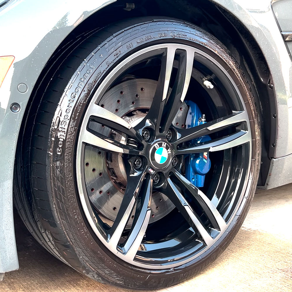 Close-up of a car wheel featuring a black alloy rim with a blue brake caliper visible behind it. The tire tread appears clean and unworn, and the car body is a shiny, light gray color.