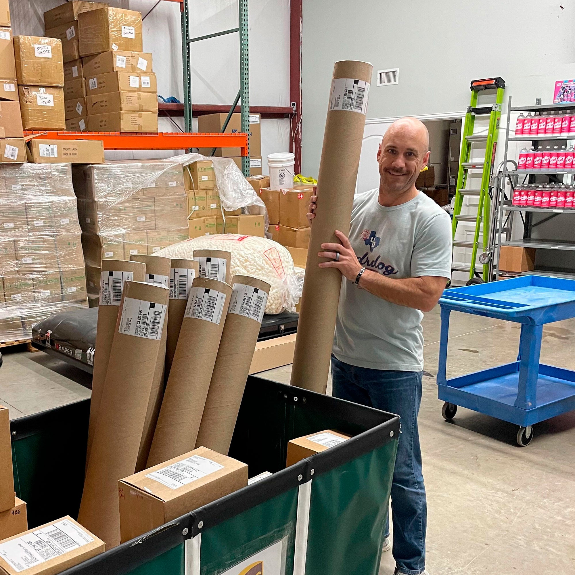 A person stands in a warehouse next to a large bin filled with long, cylindrical cardboard tubes. More boxes are stacked in the background, and a ladder is against the wall. The person is wearing a light gray T-shirt and jeans.