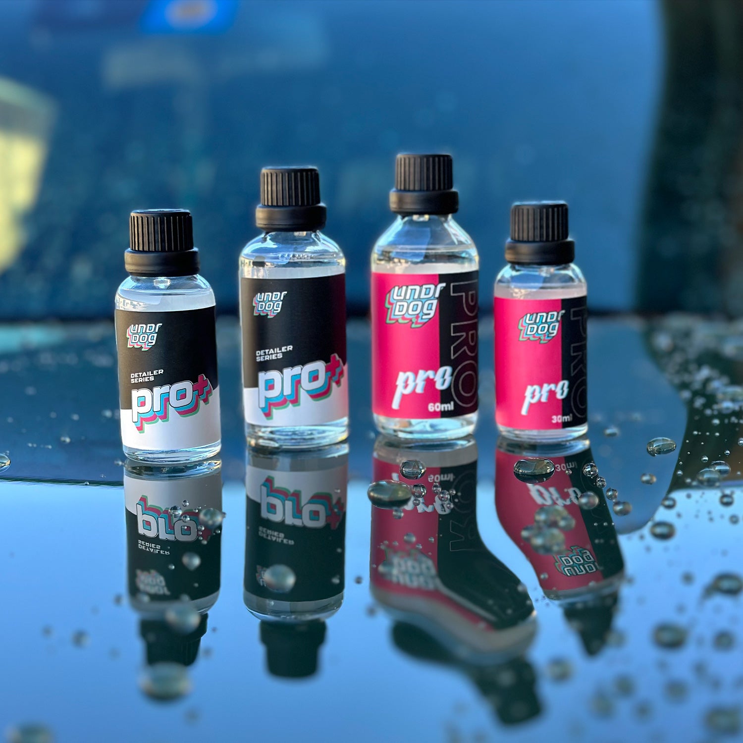 Four bottles of Undrdog Pro automotive coating are displayed on a wet car surface. The bottles, with different colored labels, vary in size and capacity. Water droplets are scattered across the reflective surface, adding texture to the scene.