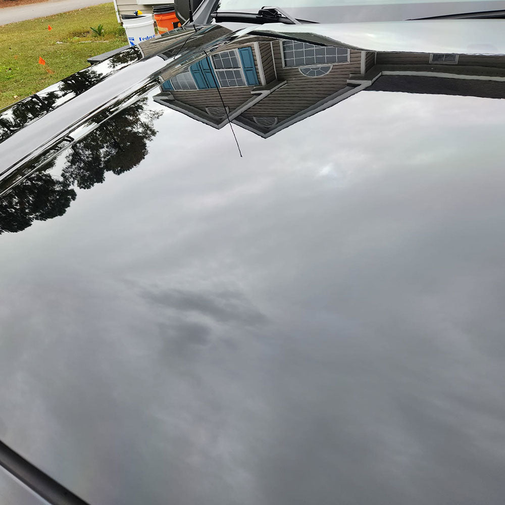 Reflection of a house and trees on the shiny surface of a black car hood. The hood displays a mirror-like finish, capturing the overcast sky above. The surroundings are slightly distorted in the reflection.