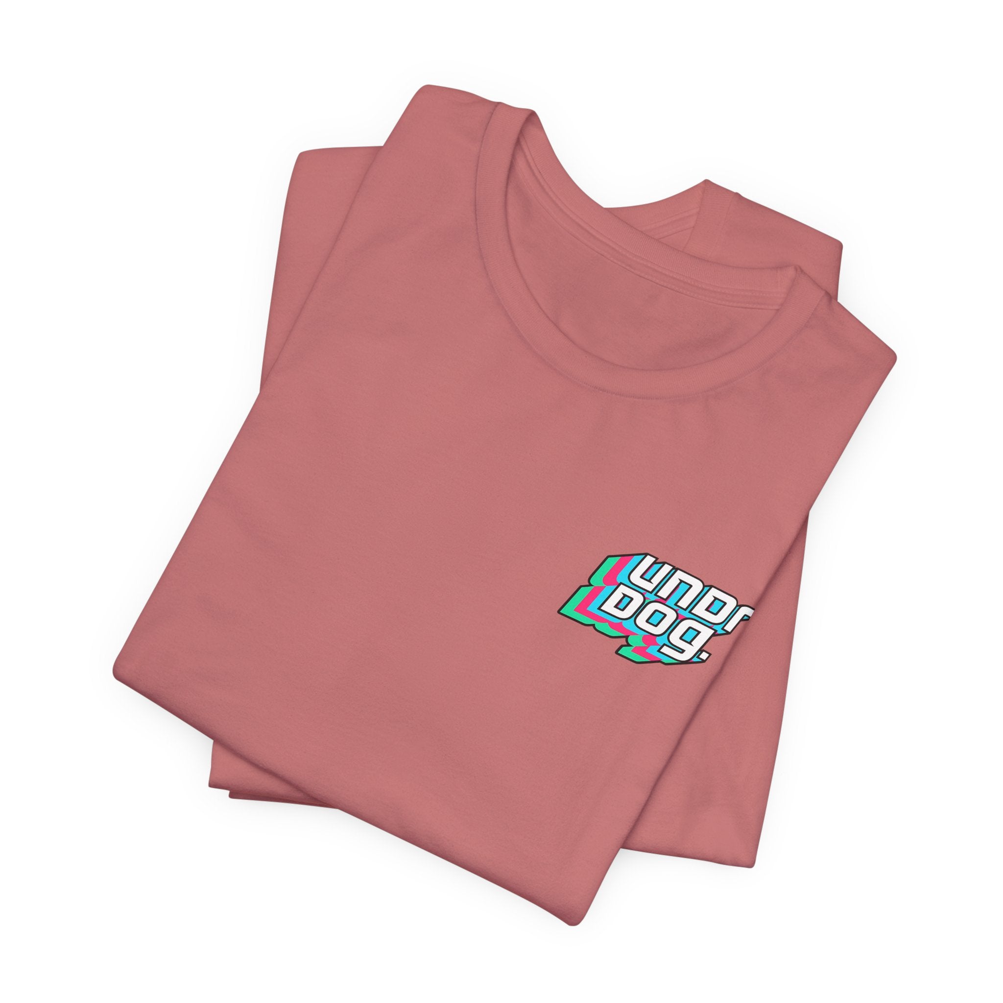 A folded mauve Unisex Jersey Short Sleeve Tee by Printify, crafted from soft Airlume combed cotton, features a small multicolored "underdog" graphic on the left chest. The shirt is displayed on a plain white background.