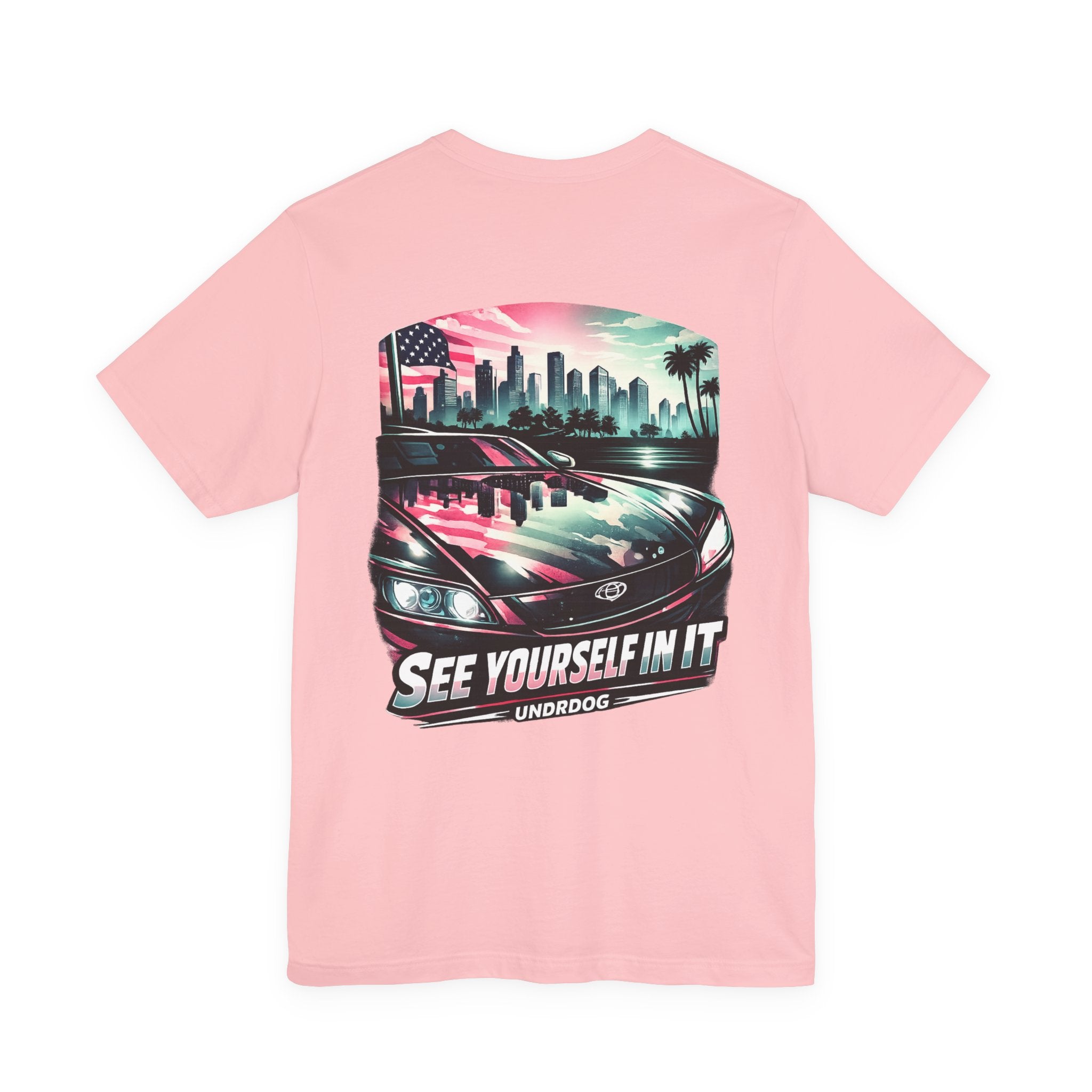 The See Yourself In It Retro Sportscar Tee by Printify features a light pink vintage car with a city skyline, palm trees, and an American flag reflection, plus "SEE YOURSELF IN IT" and "UNDRDOG" text on the back.