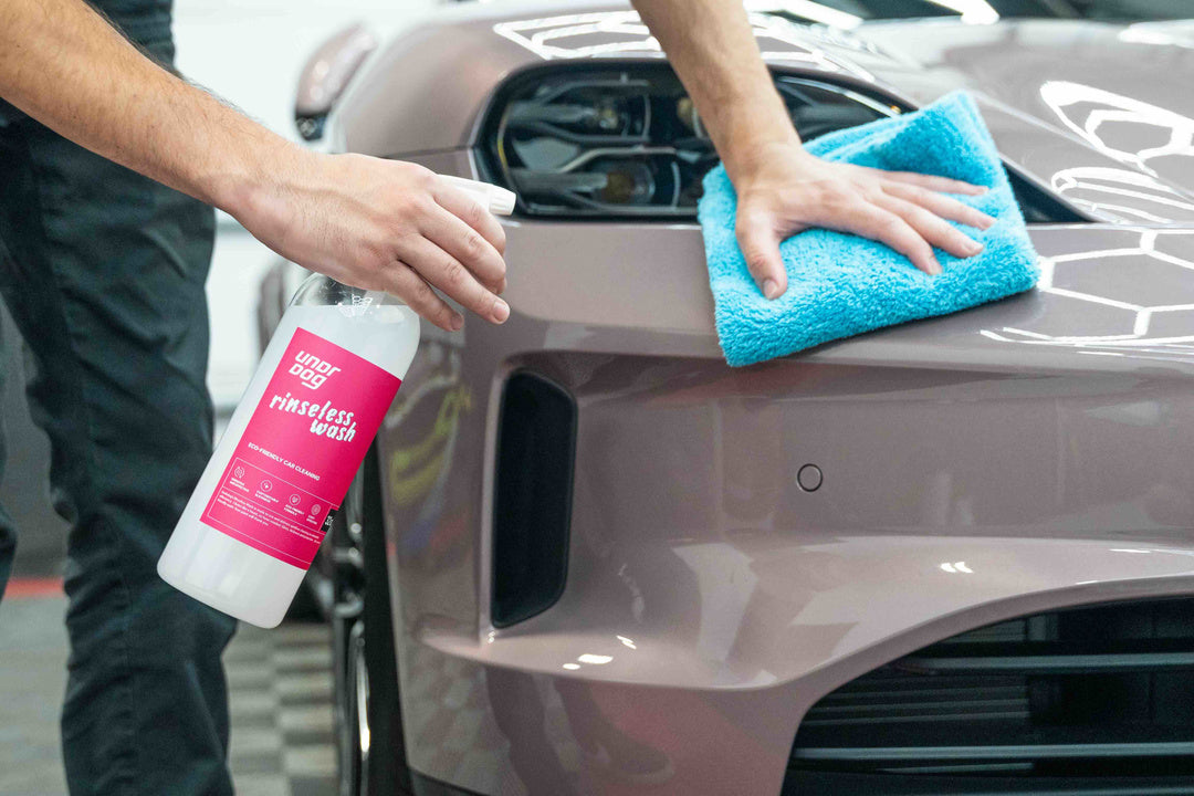 Someone cleans a mauve car's front headlight area using Undrdog Surface Products' Rinseless Wash Spray Bundle Pack and a blue microfiber cloth. A spray bottle labeled "rinse less wash" is in hand.