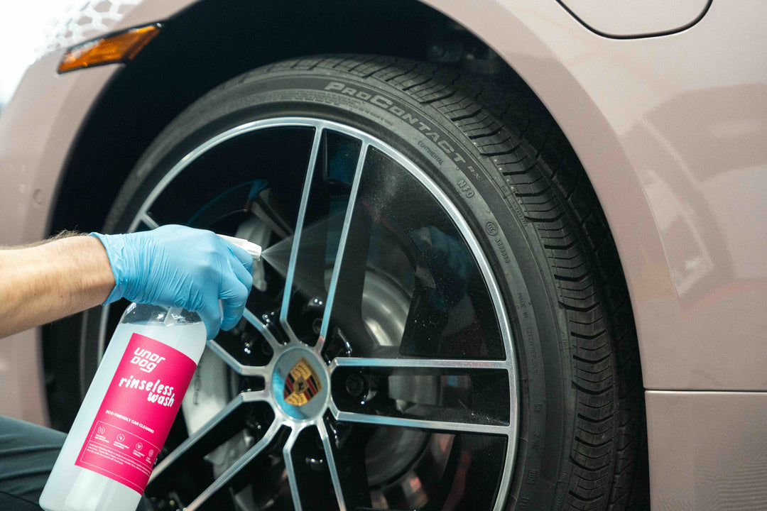 A person wearing blue gloves sprays Undrdog Surface Products Rinseless Wash Spray Bundle Pack onto a Porsche wheel, focusing on cleaning the tire and rim.