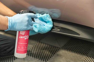 Wearing blue gloves, a person cleans a car door with a light blue microfiber cloth, holding a spray bottle of Undrdog Surface Products Rinseless Wash Spray in the other hand.