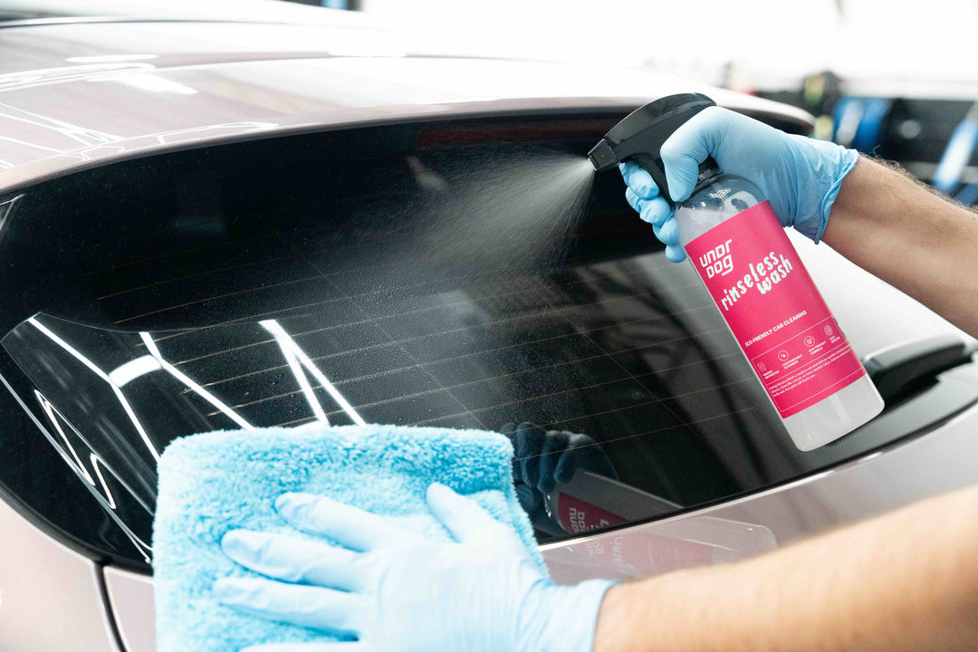 A person in blue gloves sprays Undrdog Surface Products' Rinseless Wash Spray Bundle Pack onto a car’s rear window while holding a blue microfiber cloth against the glass.