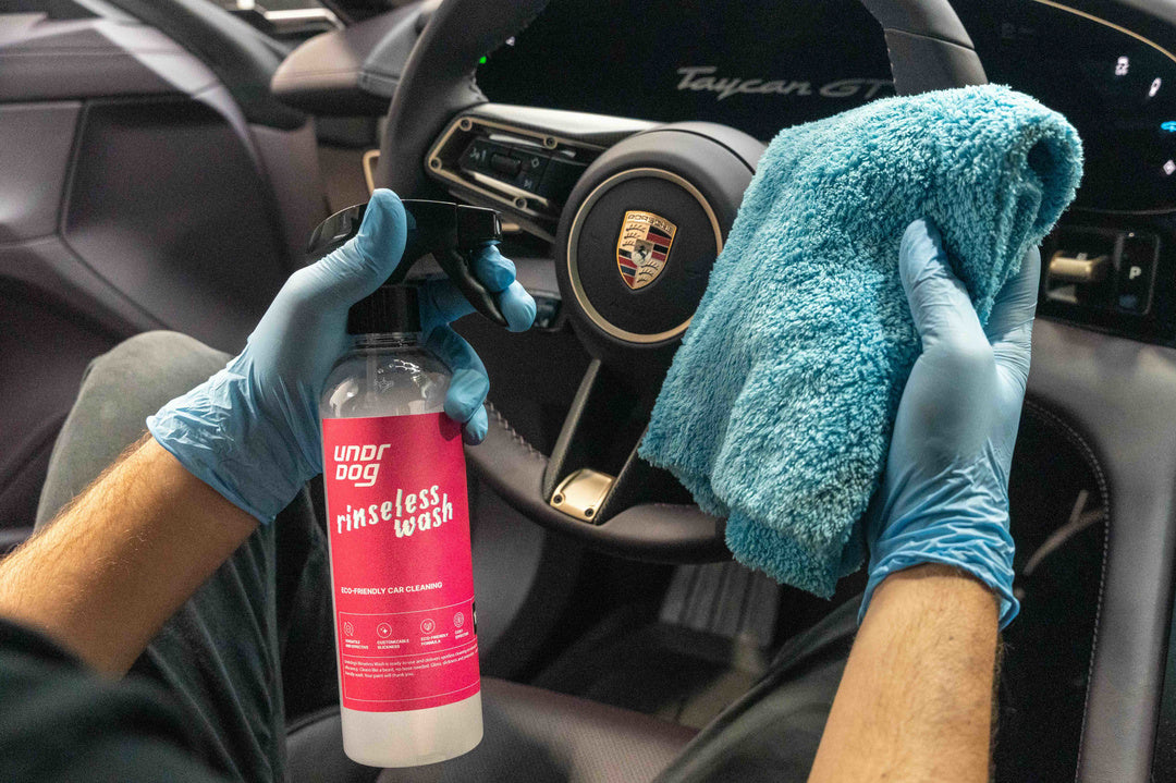 Inside a Porsche Taycan, a person in blue gloves holds Undrdog Surface Products’ Rinseless Wash Spray Bundle Pack and a blue microfiber cloth, with the steering wheel and dashboard visible.