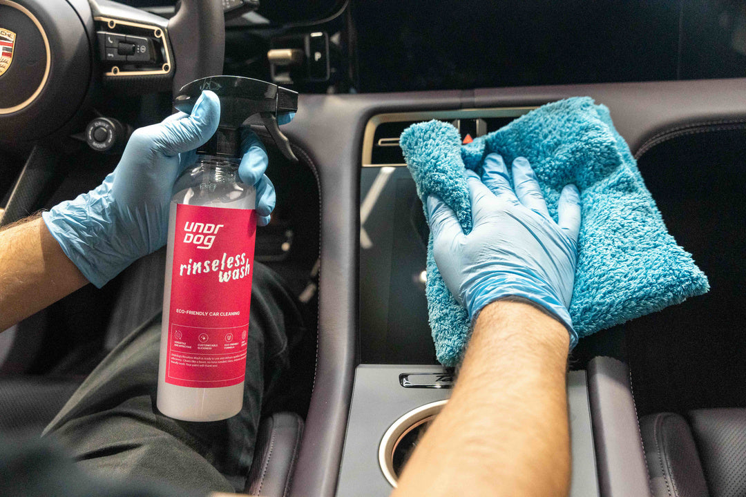 Wearing blue gloves, a person cleans a modern car’s center console—featuring a cup holder—using a blue microfiber cloth and a spray bottle of Undrdog Surface Products Rinseless Wash Spray.