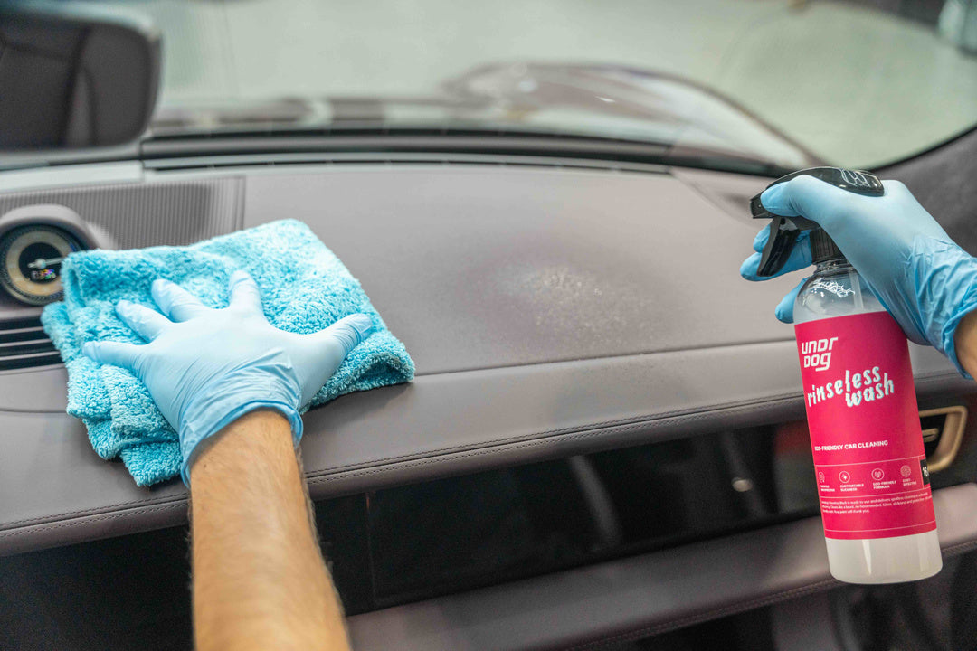 Wearing blue gloves, a person sprays Undrdog Surface Products' Rinseless Wash Spray Bundle Pack onto a car dashboard and wipes it clean with a blue microfiber cloth.
