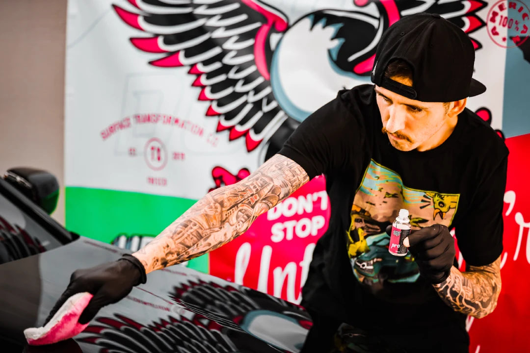 A man in a backward cap polishes a black car with a pink cloth, holding Undrdog Surface Products’ HCC - Hybrid Ceramic Composite. He wears black gloves, a graphic tee, and stands before a colorful winged banner.